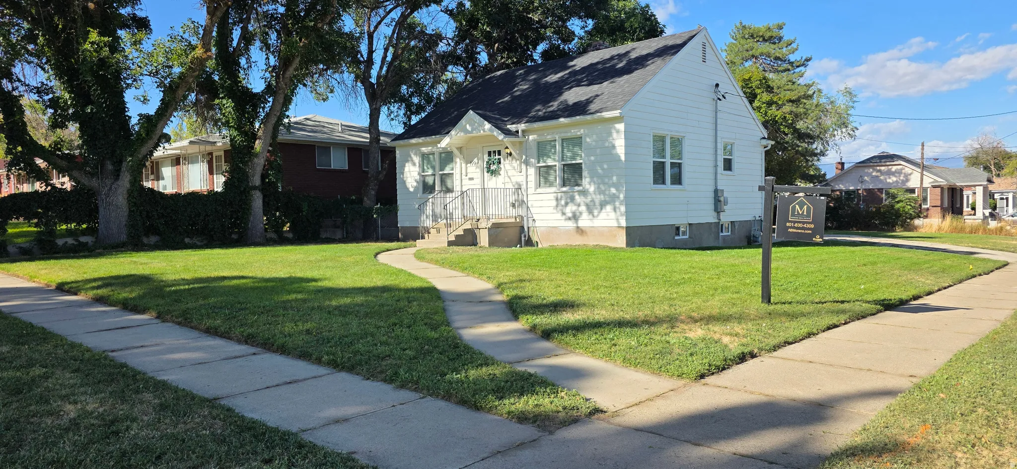 White siding exterior with a dark shingle roof, established trees, and a concrete walkway