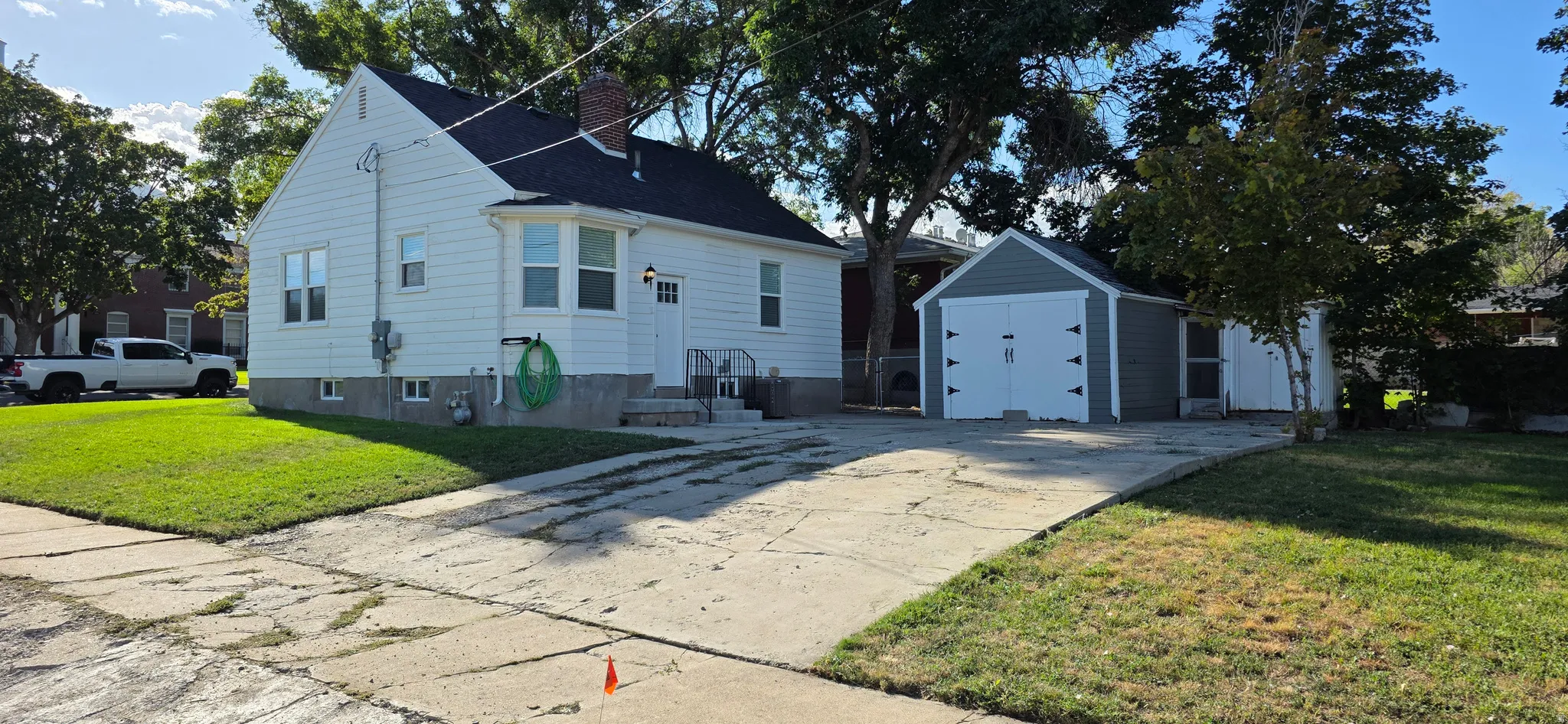 White siding exterior with a dark shingle roof, concrete driveway, detached garage with white doors, and mature trees