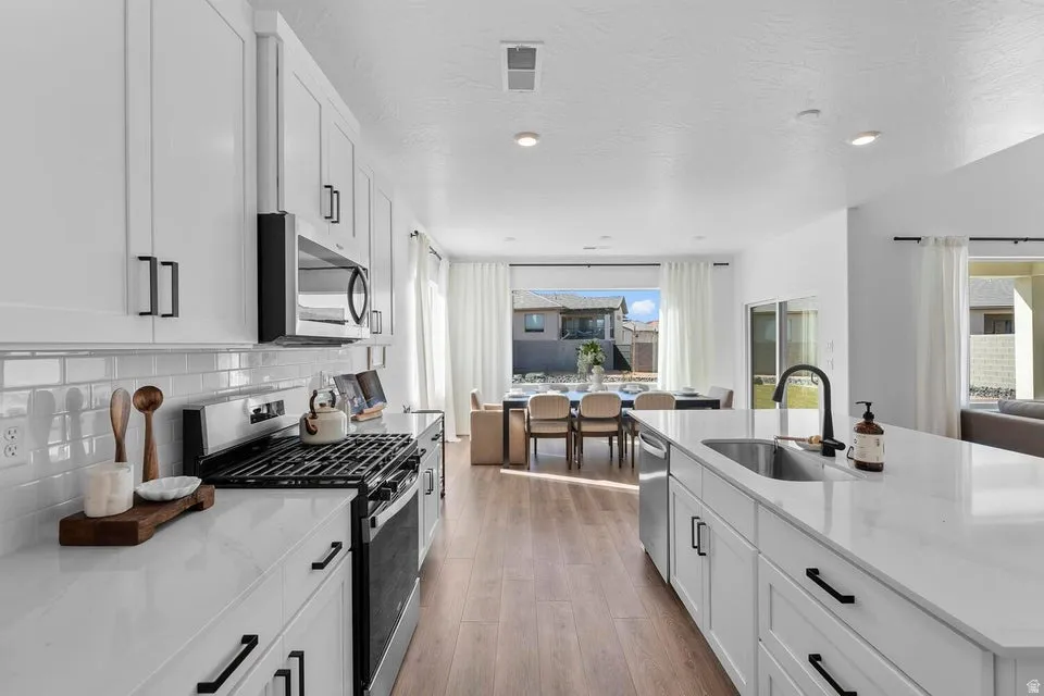 Open-concept kitchen featuring white cabinetry, stainless steel appliances, a white subway tile backsplash, a kitchen island with an undermount sink, and wood-finish flooring