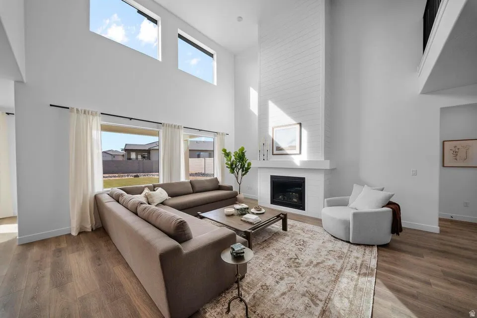 Two-story living area with high ceilings, multiple large windows, wood-finish flooring, a white brick-finish fireplace, and an upper-level balcony