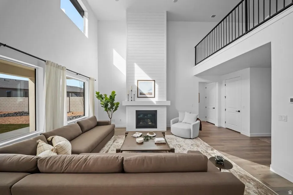 Two-story living area with high ceilings, wood-finish flooring, a contemporary fireplace with shiplap surround, large windows, and an upper-level balcony with metal railings