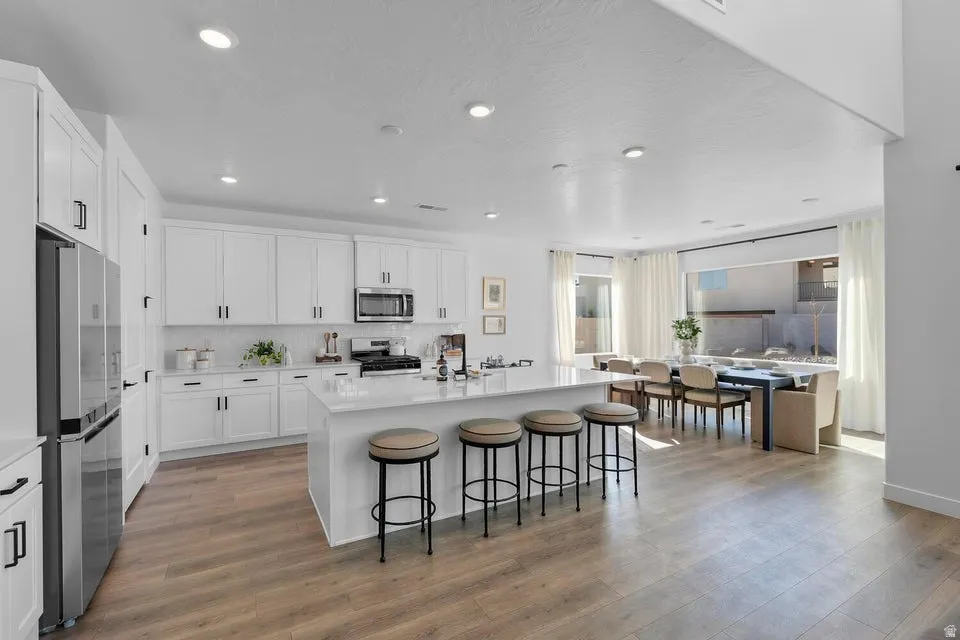 White shaker cabinetry, stainless steel appliances, a large center island with seating, wood-finish flooring, and recessed lighting