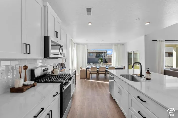 Open-concept kitchen featuring white cabinetry, stainless steel appliances, a white subway tile backsplash, a kitchen island with an undermount sink, and wood-finish flooring