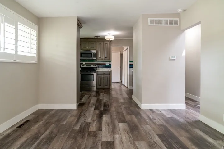View from the living room into the kitchen, LVP flooring, white plantation shutters, neutral wall tones, stainless steel appliances, and ceiling-mounted lighting.