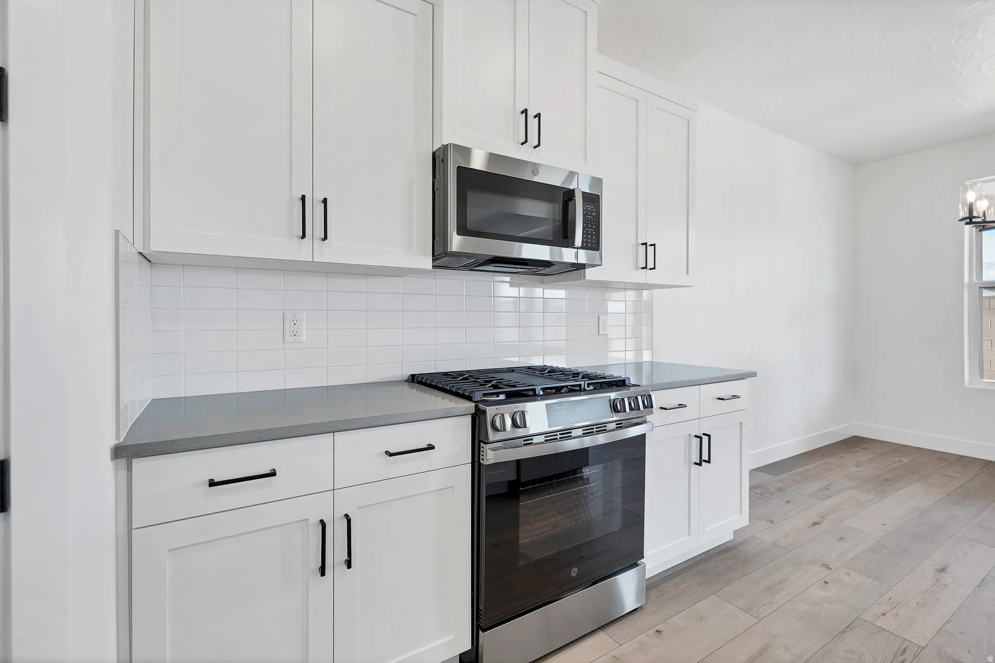 White shaker cabinetry with matte black hardware, stainless steel range and microwave, light gray countertops, subway tile backsplash, and wood-finish flooring
