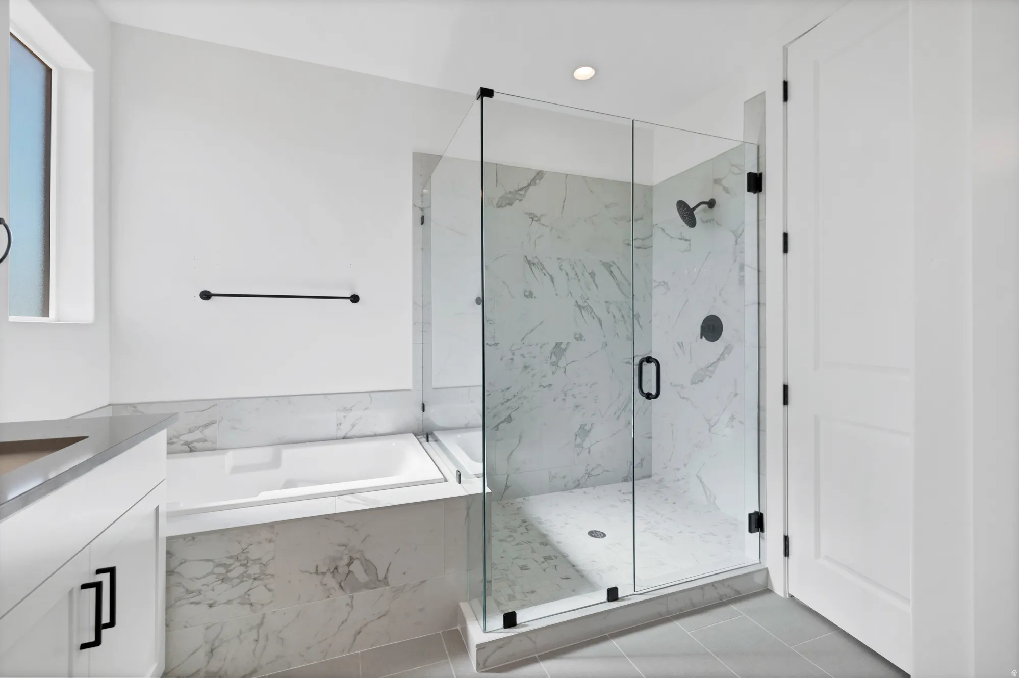 Enclosed glass shower with dark fixtures and marbled wall, freestanding bathtub, modern vanity with dark hardware, and light-colored tile flooring