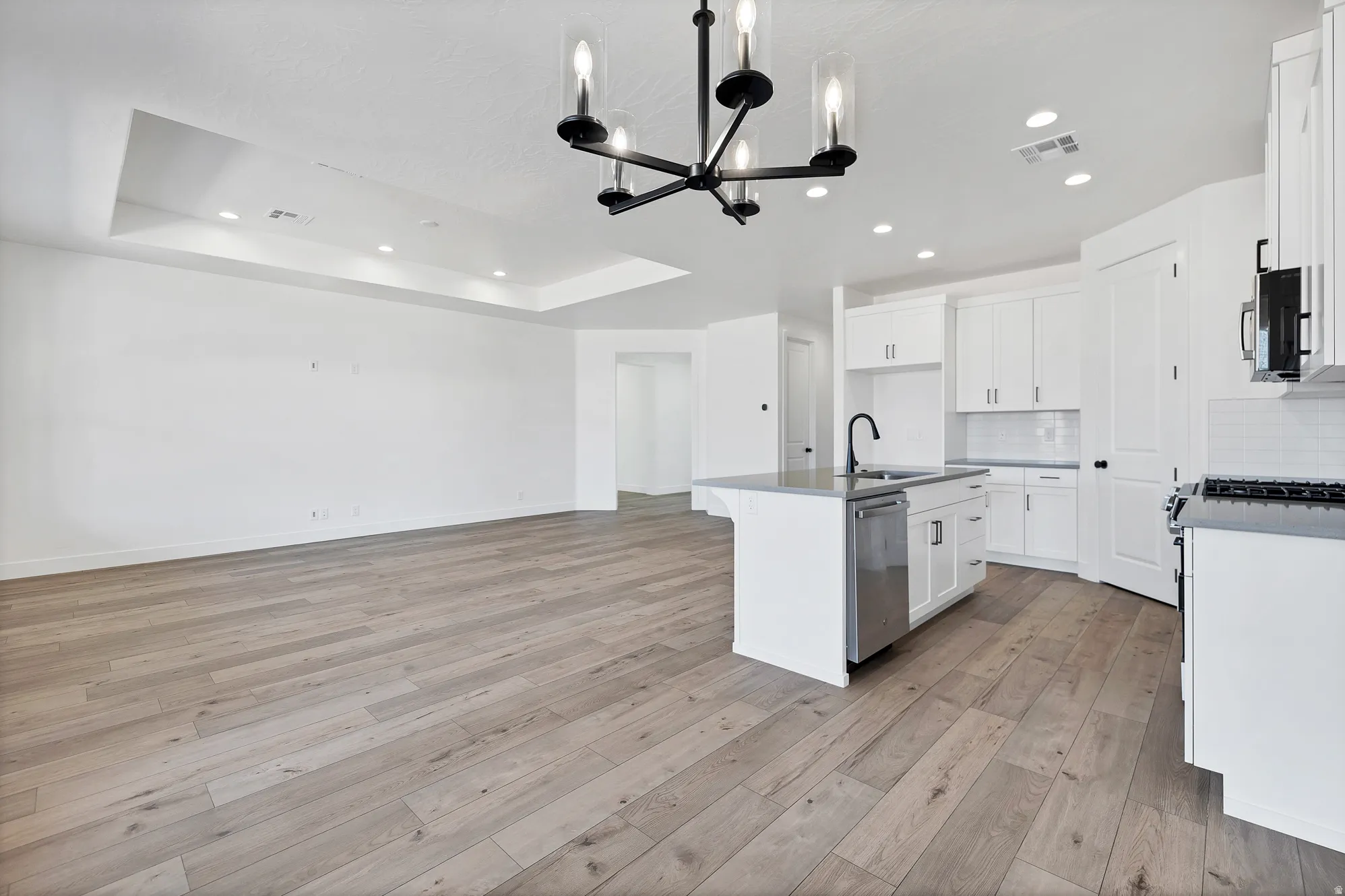 Expansive open-concept space featuring wood-finish flooring, a contemporary black chandelier, recessed lighting, a tray ceiling, and a kitchen island with an undermount sink