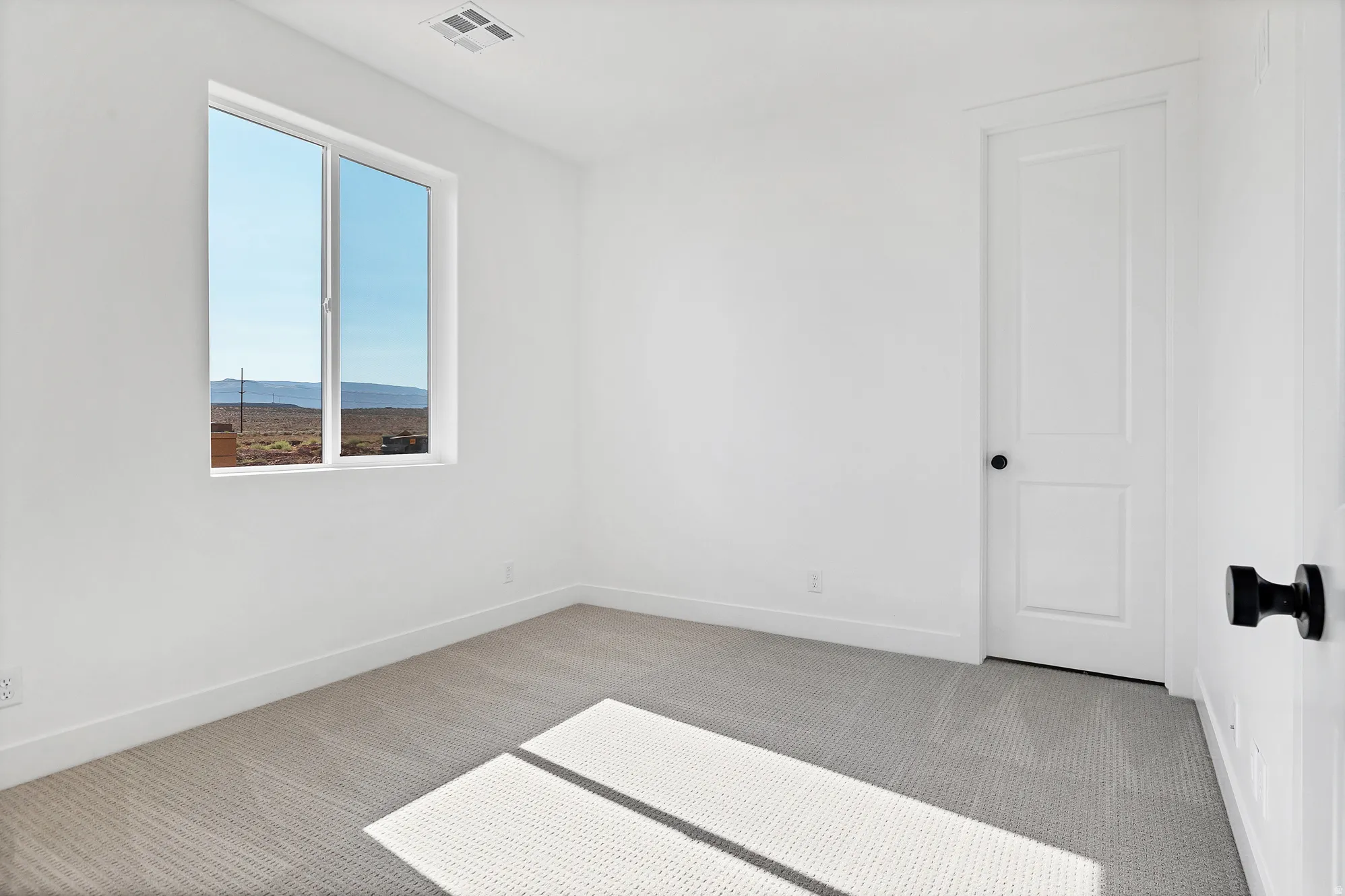 White walls with light gray patterned carpeting, a single window with a desert landscape, a solid white door with a black knob, and a ceiling vent