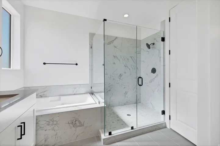 Enclosed glass shower with dark fixtures and marbled wall, freestanding bathtub, modern vanity with dark hardware, and light-colored tile flooring