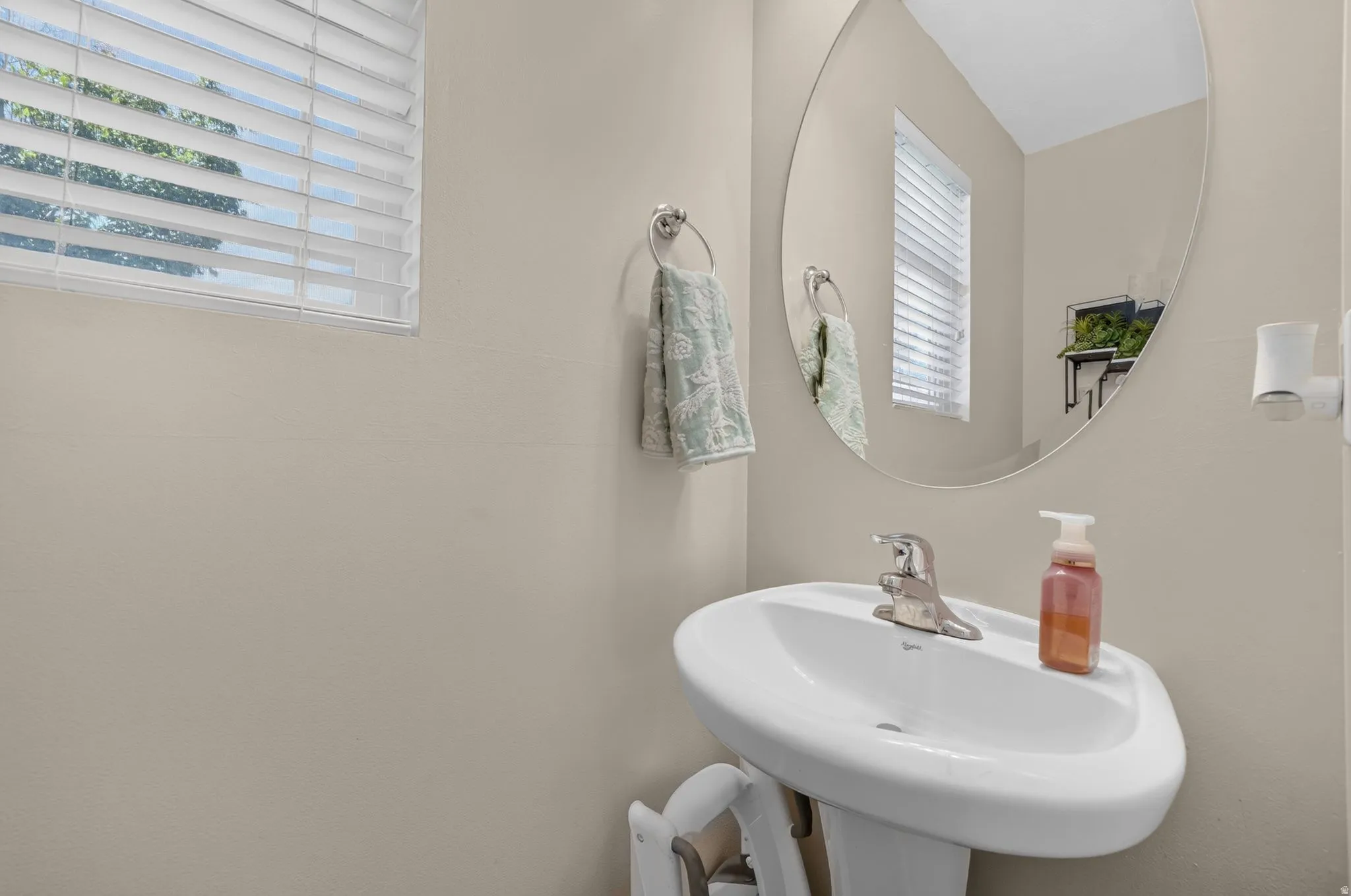 Pedestal sink with chrome faucet, oval wall mirror, and window with blinds