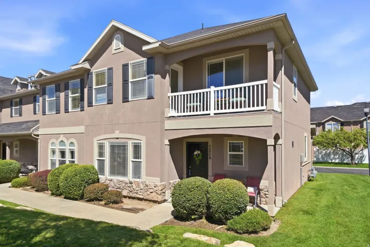 Stucco exterior with stone accents, covered entry, second-story balcony with white railing, manicured shrubs, and a well-maintained lawn