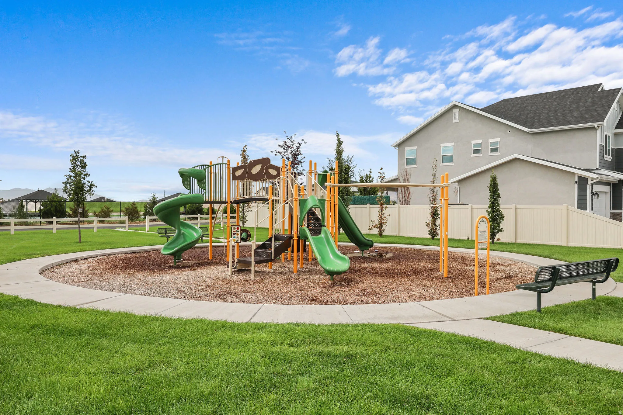Community playground with multiple slides, swing set, and a bench, surrounded by a paved walking path and expansive green space