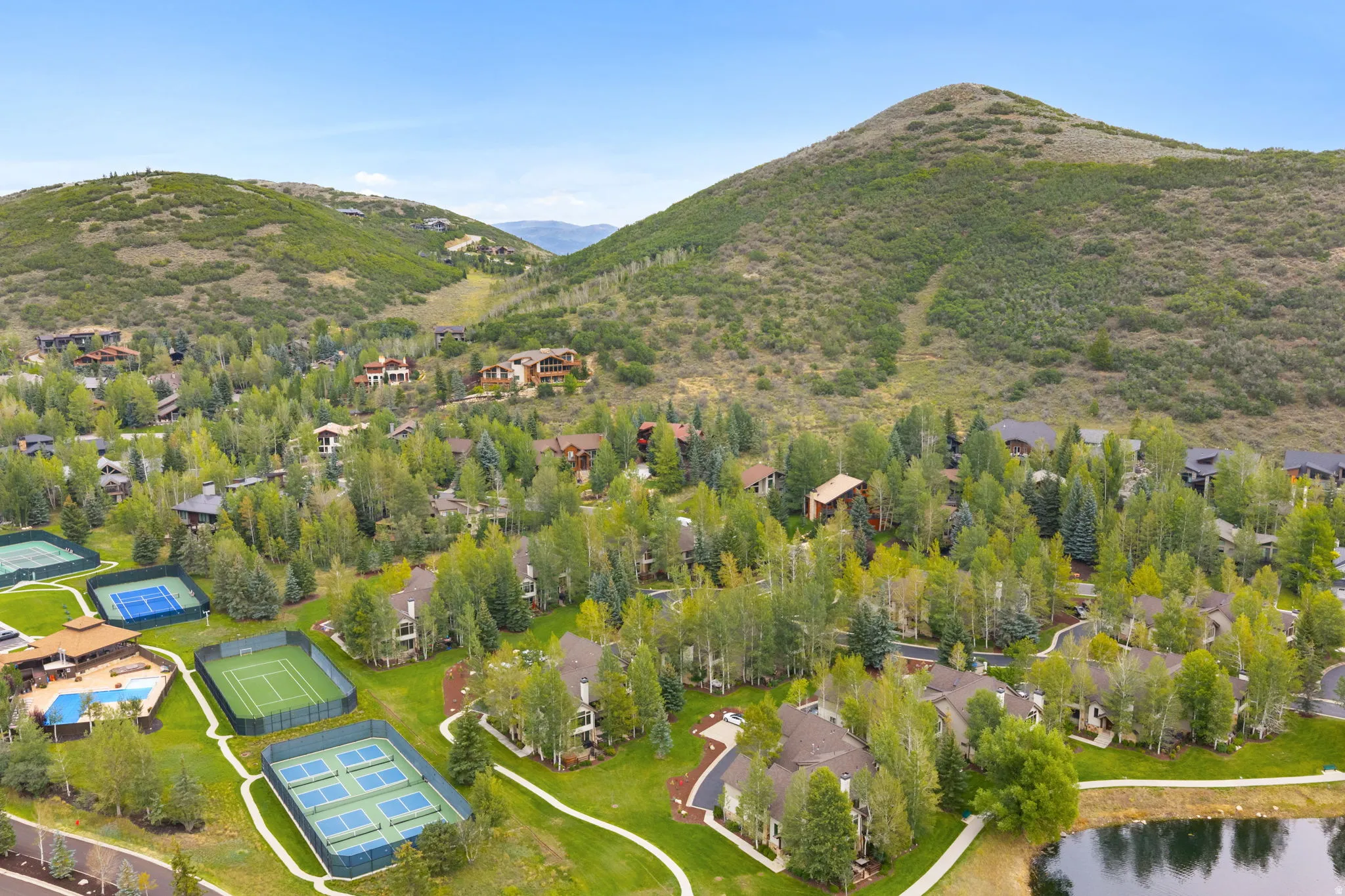 Aerial perspective showcasing a community nestled among lush trees, featuring multiple tennis courts, a swimming pool, and a tranquil pond, all set against a mountainous backdrop