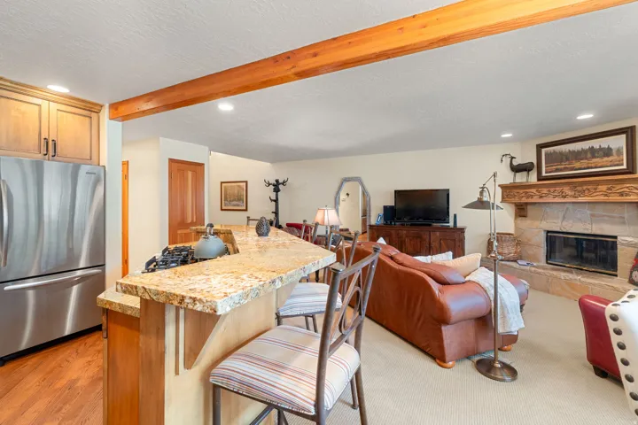Open-concept living space featuring a stone-clad fireplace with carved wood mantel, wood-finish flooring, a kitchen island with stone countertops, and stainless steel appliances