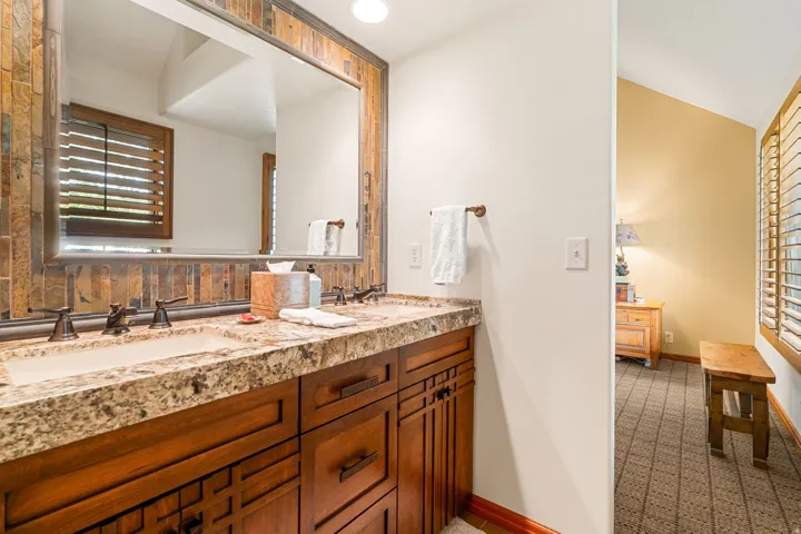 Dual undermount sinks with bronze-finish fixtures, a large mirror framed by natural stone tile, and wood-finish cabinetry