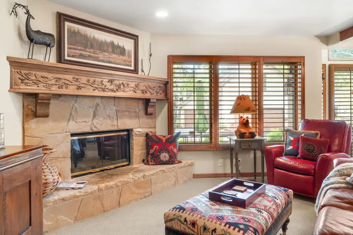 Stone fireplace with a carved wood mantel, carpeted flooring, and wood-framed windows with plantation shutters