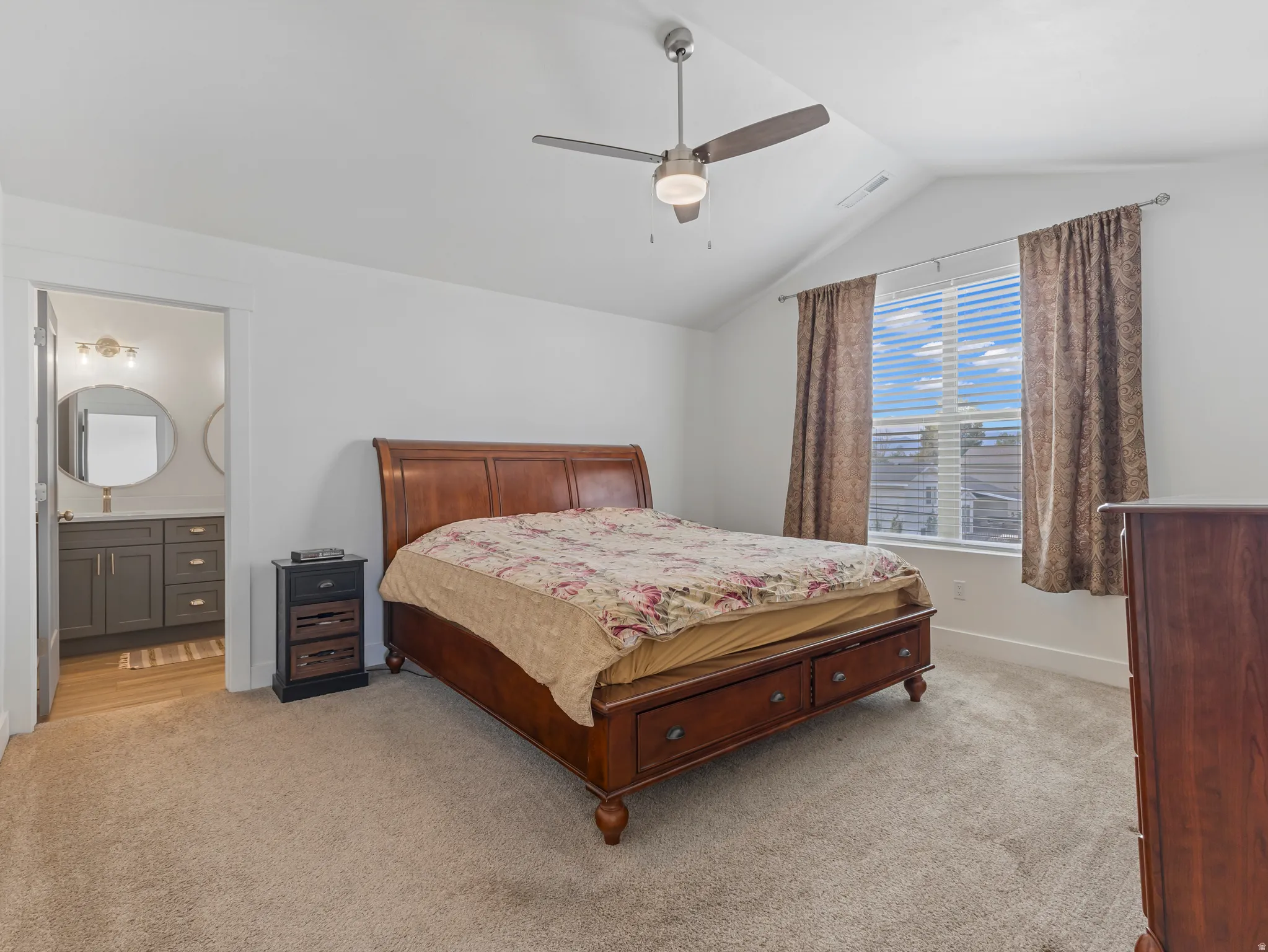 Vaulted ceiling with a ceiling fan, broad window with horizontal blinds, carpeting, and an adjoining bathroom featuring a vanity with two circular mirrors, a single basin, and wood-finish flooring