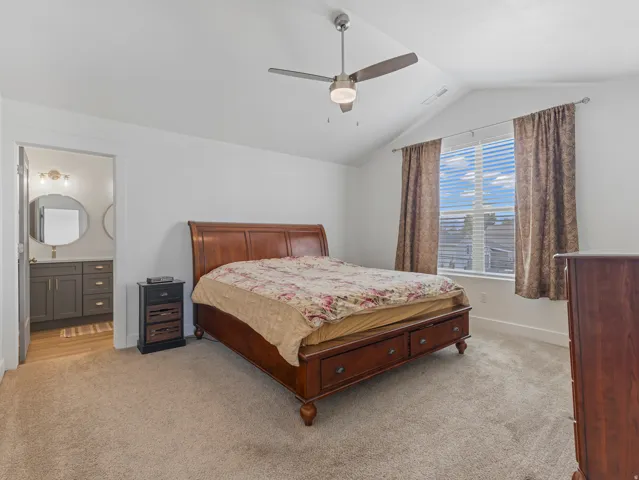 Vaulted ceiling with a ceiling fan, broad window with horizontal blinds, carpeting, and an adjoining bathroom featuring a vanity with two circular mirrors, a single basin, and wood-finish flooring