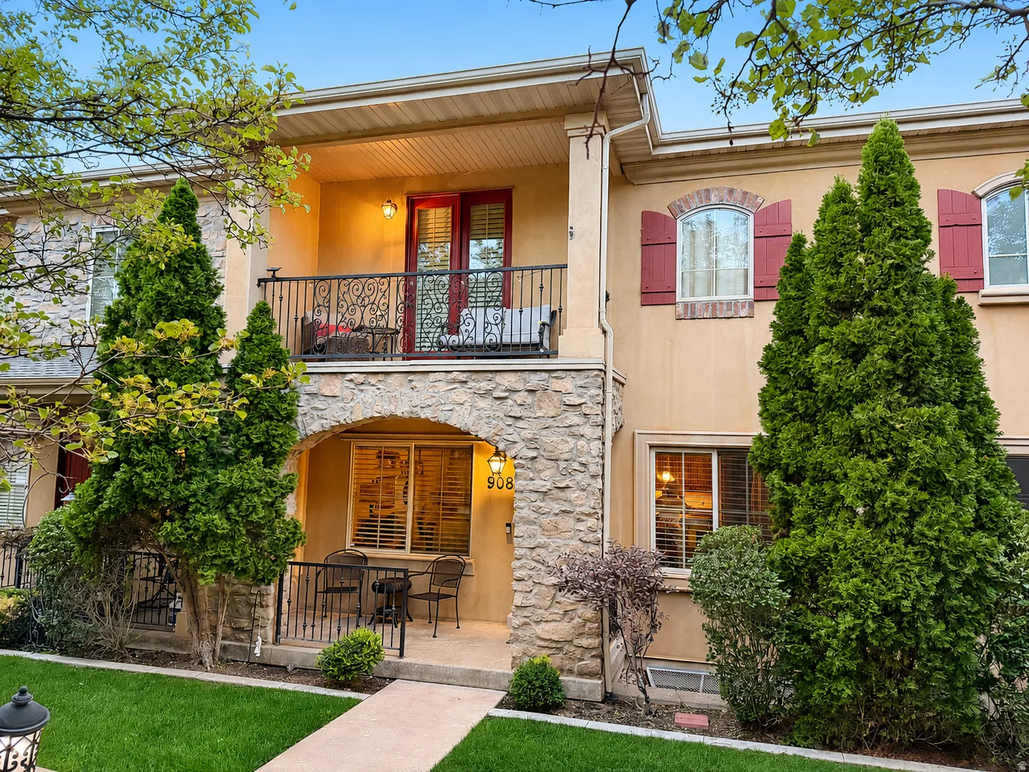 Stucco exterior with stone archway, second-story balcony with wrought iron railing, arched windows with red shutters, and manicured landscaping
