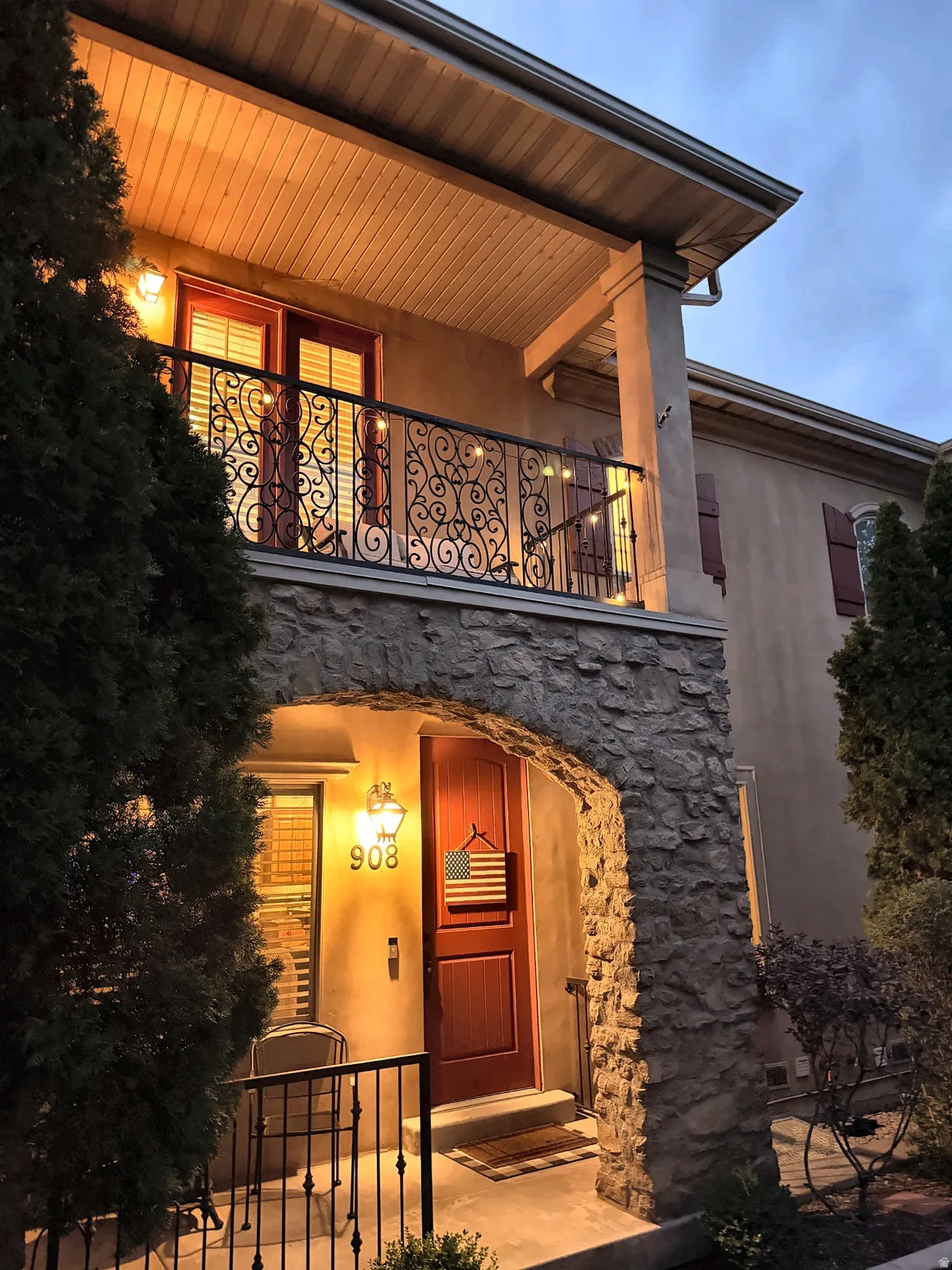 Stone archway entryway with a rich wood-finish door, decorative wall sconce, and an upstairs balcony featuring an ornate iron railing