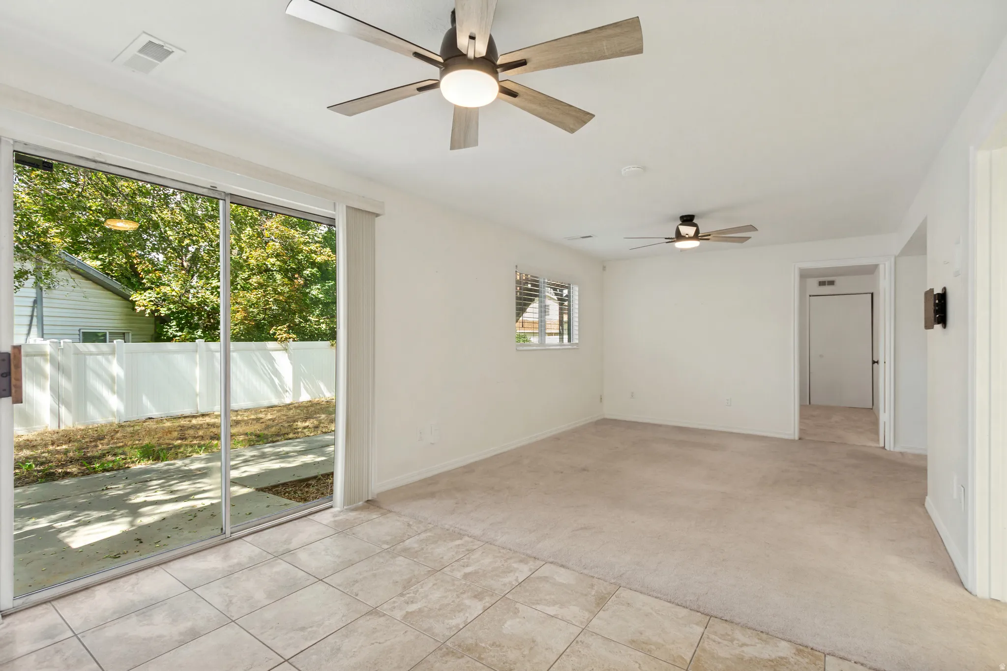 Sliding glass doors opening to the exterior, two ceiling fans with integrated lighting, a rectangular window with horizontal blinds, and a combination of carpet and tiled flooring