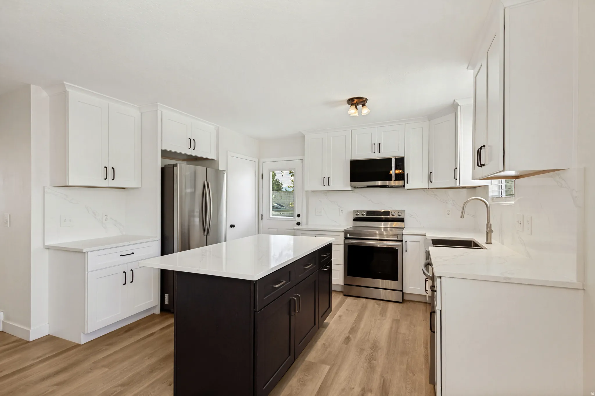 White shaker cabinetry, stainless steel appliances, wood-finish flooring, central island with dark cabinetry, and white countertops