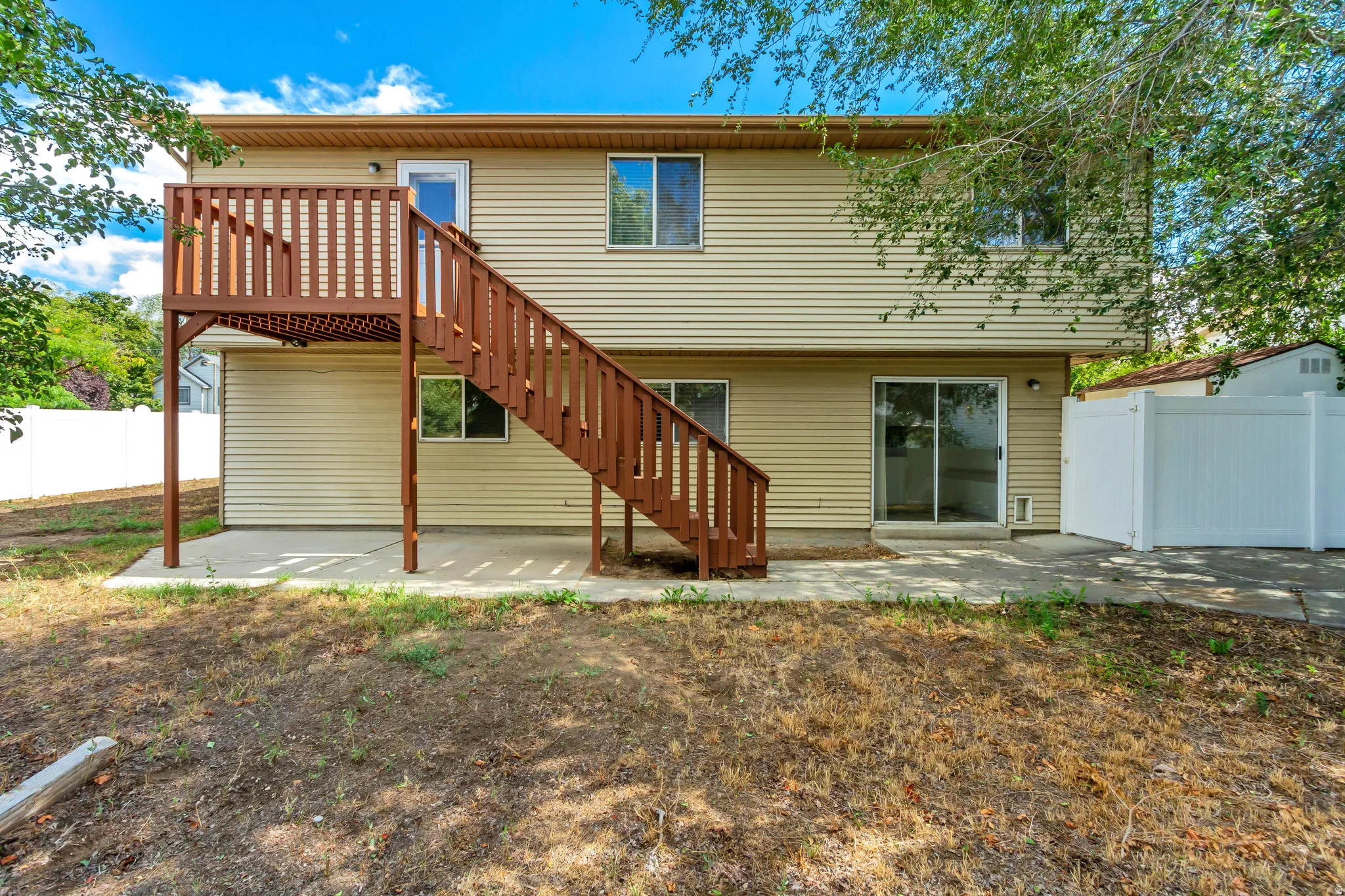 Rear exterior featuring a second-story wood deck with stairs, beige horizontal siding, a sliding glass door, multiple windows, and a white privacy fence