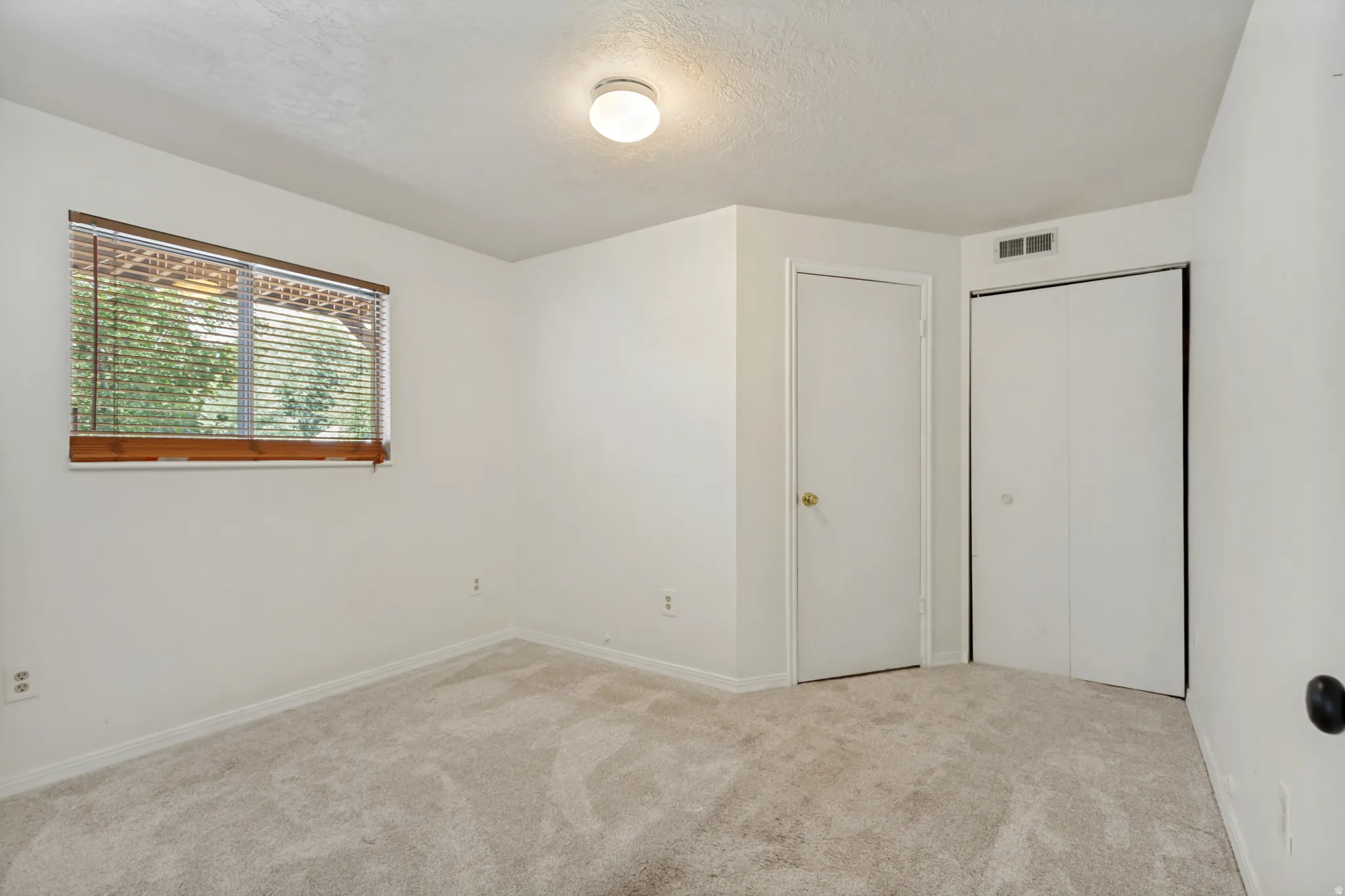 Neutral-toned carpet flooring, white walls, wood-framed window with blinds, white flush-mount ceiling light, and white bi-fold closet doors