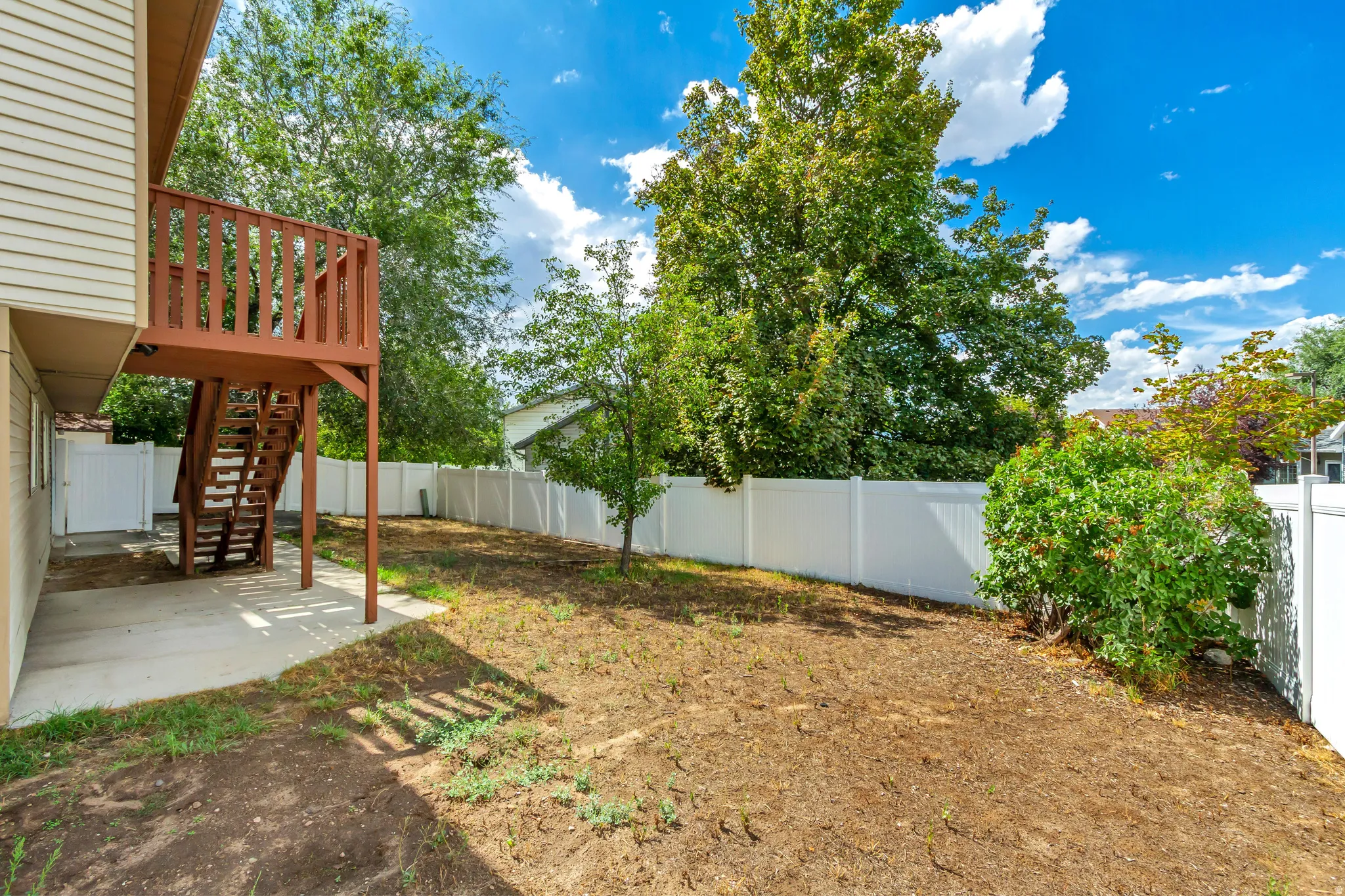 Rear exterior with a wood-stained deck and stairs, concrete patio, and white privacy fencing
