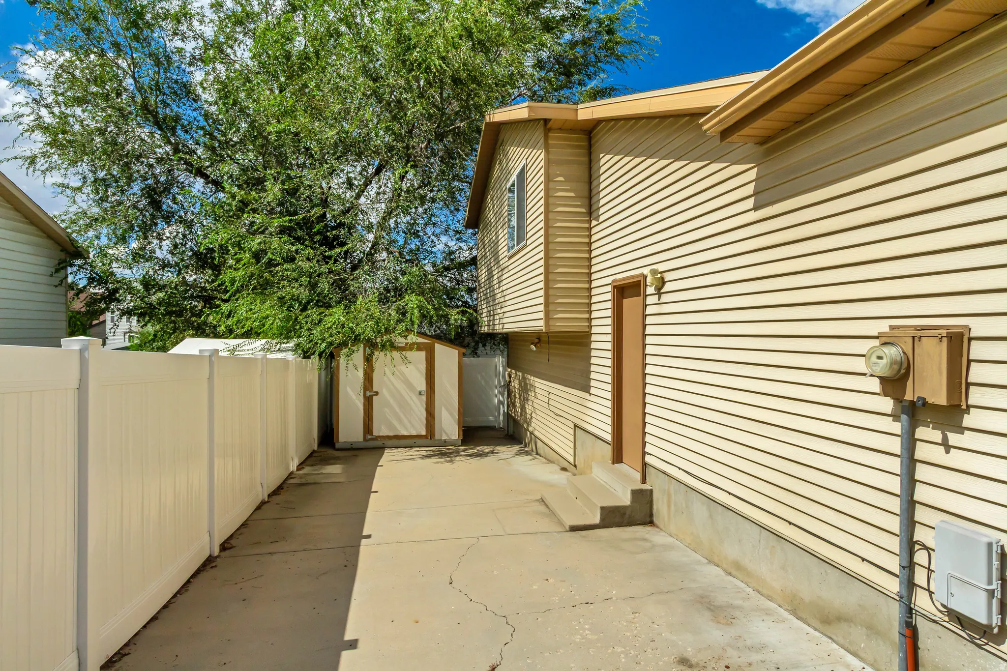 Cream-colored vinyl siding exterior with a concrete pathway, privacy fencing, and an on-site storage shed