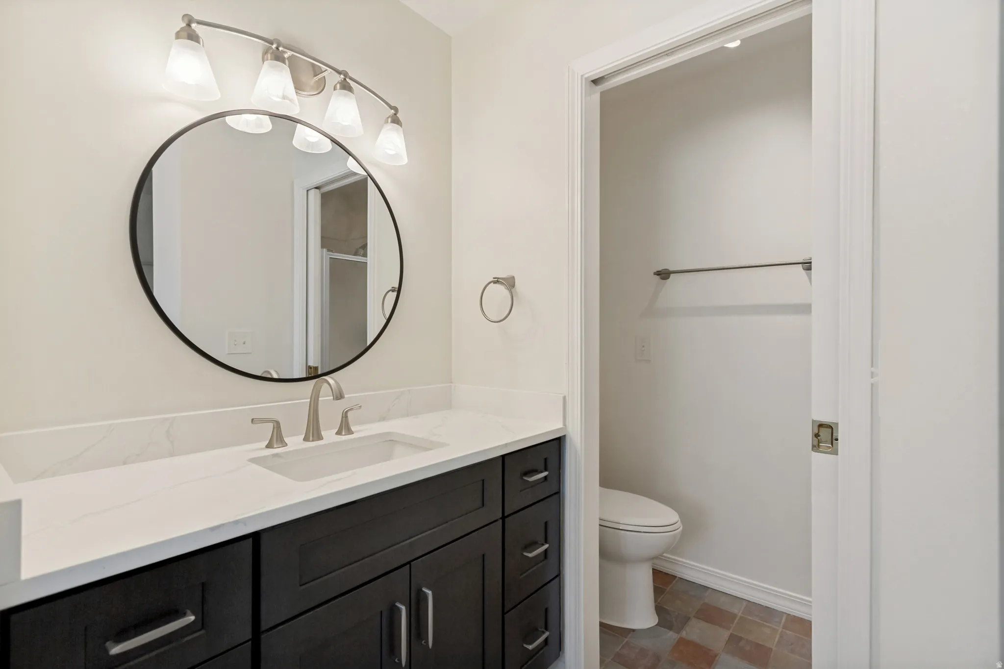 Dark wood-finish vanity with brushed nickel hardware, white countertop, rectangular undermount sink with brushed nickel faucet, round black-framed mirror, and a five-light vanity fixture