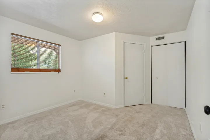 Neutral-toned carpet flooring, white walls, wood-framed window with blinds, white flush-mount ceiling light, and white bi-fold closet doors