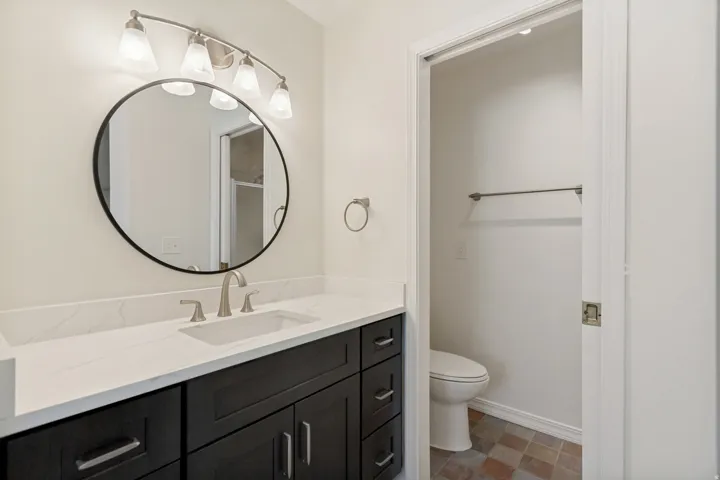 Dark wood-finish vanity with brushed nickel hardware, white countertop, rectangular undermount sink with brushed nickel faucet, round black-framed mirror, and a five-light vanity fixture