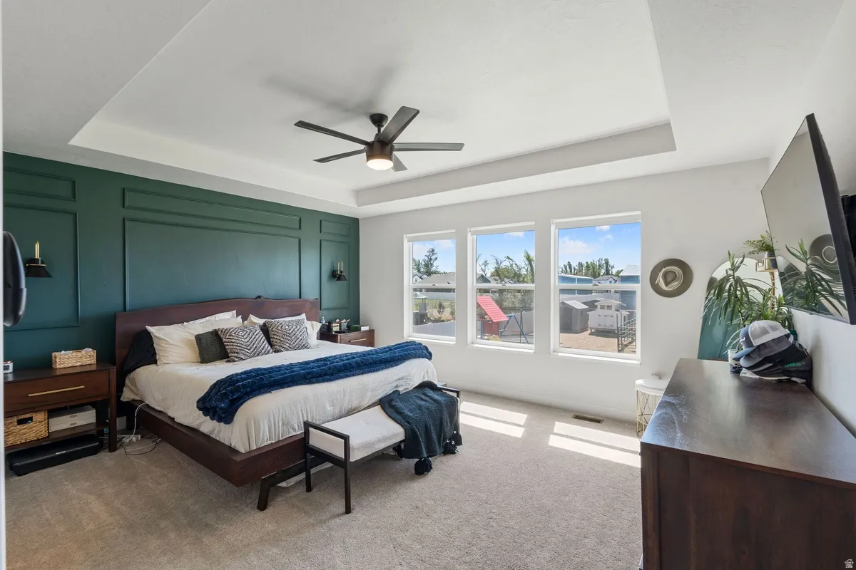 Tray ceiling with a ceiling fan, accent wall with panel molding, three windows with white trim, and light-toned carpeting