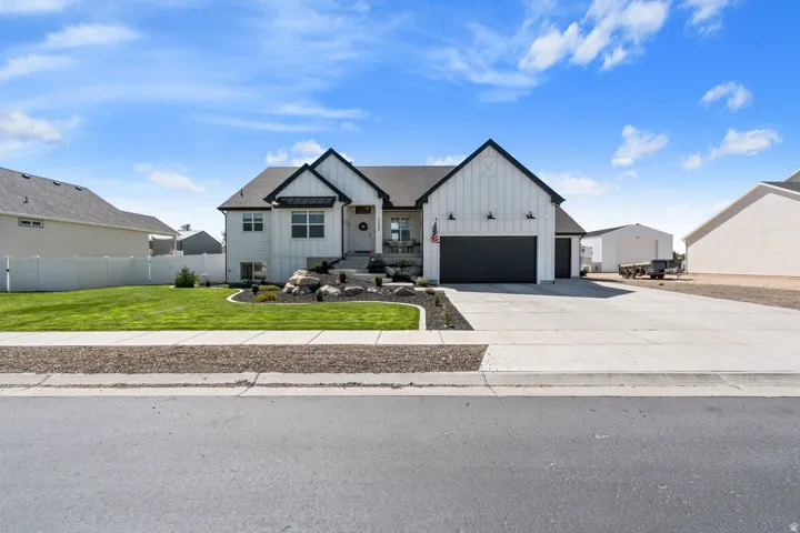 Modern farmhouse exterior with a dark-pitched roof, white vertical and horizontal siding, an attached two-car garage with a dark door, a concrete driveway, and a landscaped rock garden