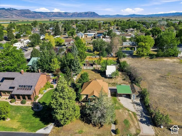 Residential property with a lush green lawn, mature trees, and a visible carport, set against a backdrop of distant mountains and open terrain