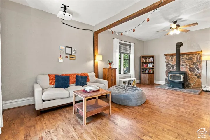 Warm wood-finish flooring, exposed ceiling beam, stone-clad wood-burning stove, and a ceiling fan with integrated lighting
