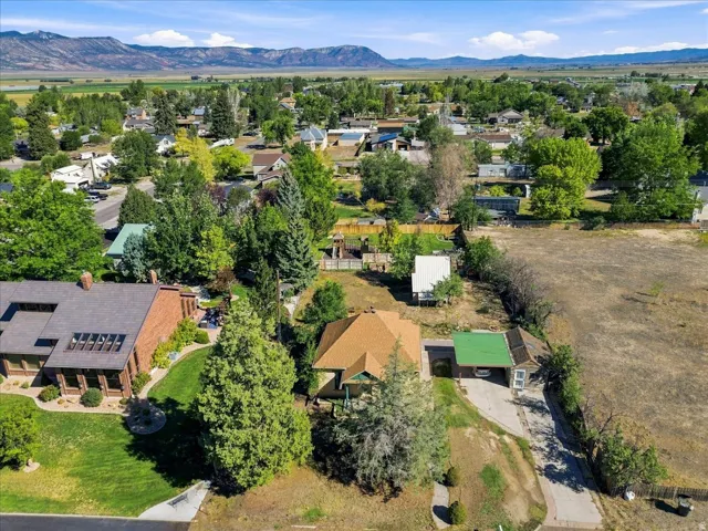Residential property with a lush green lawn, mature trees, and a visible carport, set against a backdrop of distant mountains and open terrain