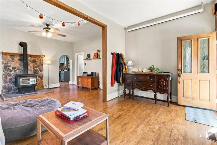 Wood-finish flooring throughout, featuring a stone-clad fireplace with a wood-burning stove, a ceiling fan, and a wood entry door with decorative glass inserts