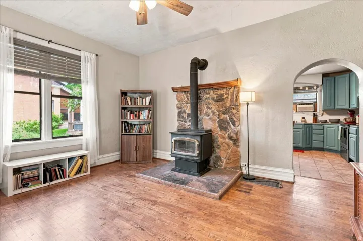 Hardwood-finish flooring, stone-faced wood-burning stove, arched doorway, ceiling fan, and large window with blinds