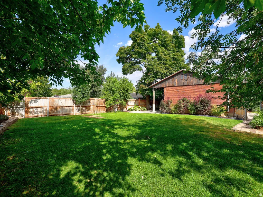 Side A fully fenced private back yard. Expansive green lawn, wood-paneled privacy fence, mature trees, brick-sided structure, and stone pathway