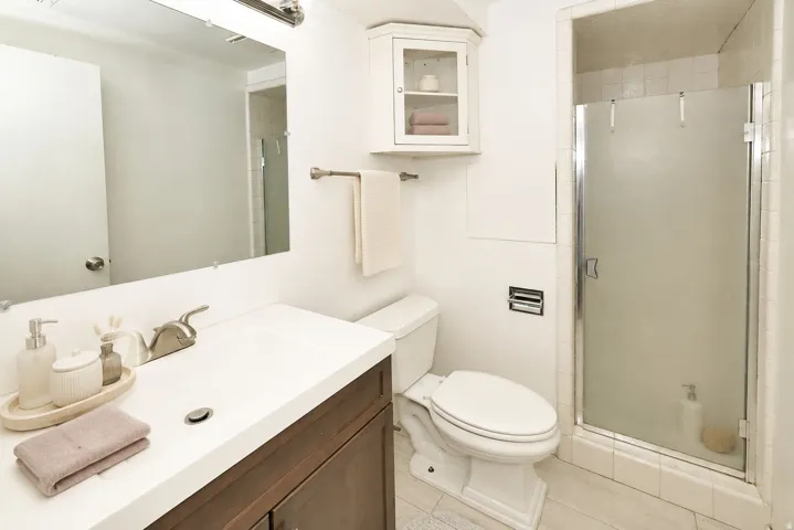 Side B White vanity with integrated sink, brushed nickel faucet, wall-mounted mirror, white wall-mounted corner cabinet, and a glass-enclosed shower