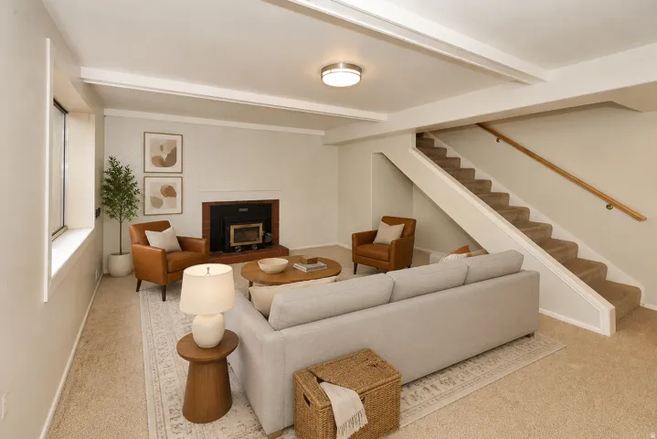 Side A basement Living area with exposed ceiling beams, carpeted flooring, a fireplace with a dark surround, and a carpeted staircase with a wood handrail