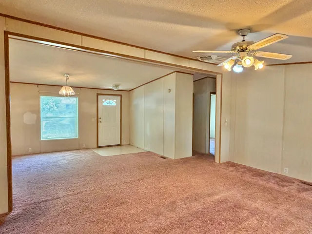 Expansive room with neutral walls, carpeted flooring, a ceiling fan with integrated lighting, and a white entry door with decorative glass