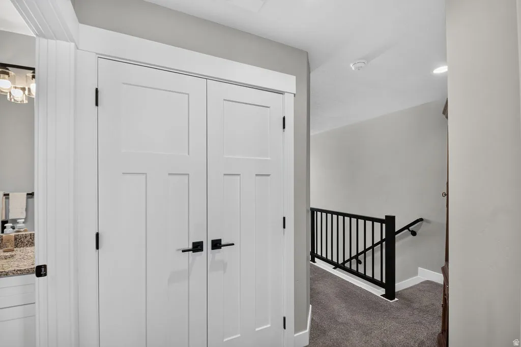 Hallway featuring white bi-fold doors with matte black hardware, a black metal staircase railing, and grey carpet flooring