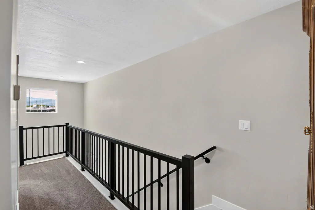 Carpeted landing featuring a black metal railing, white walls, and a window with blinds