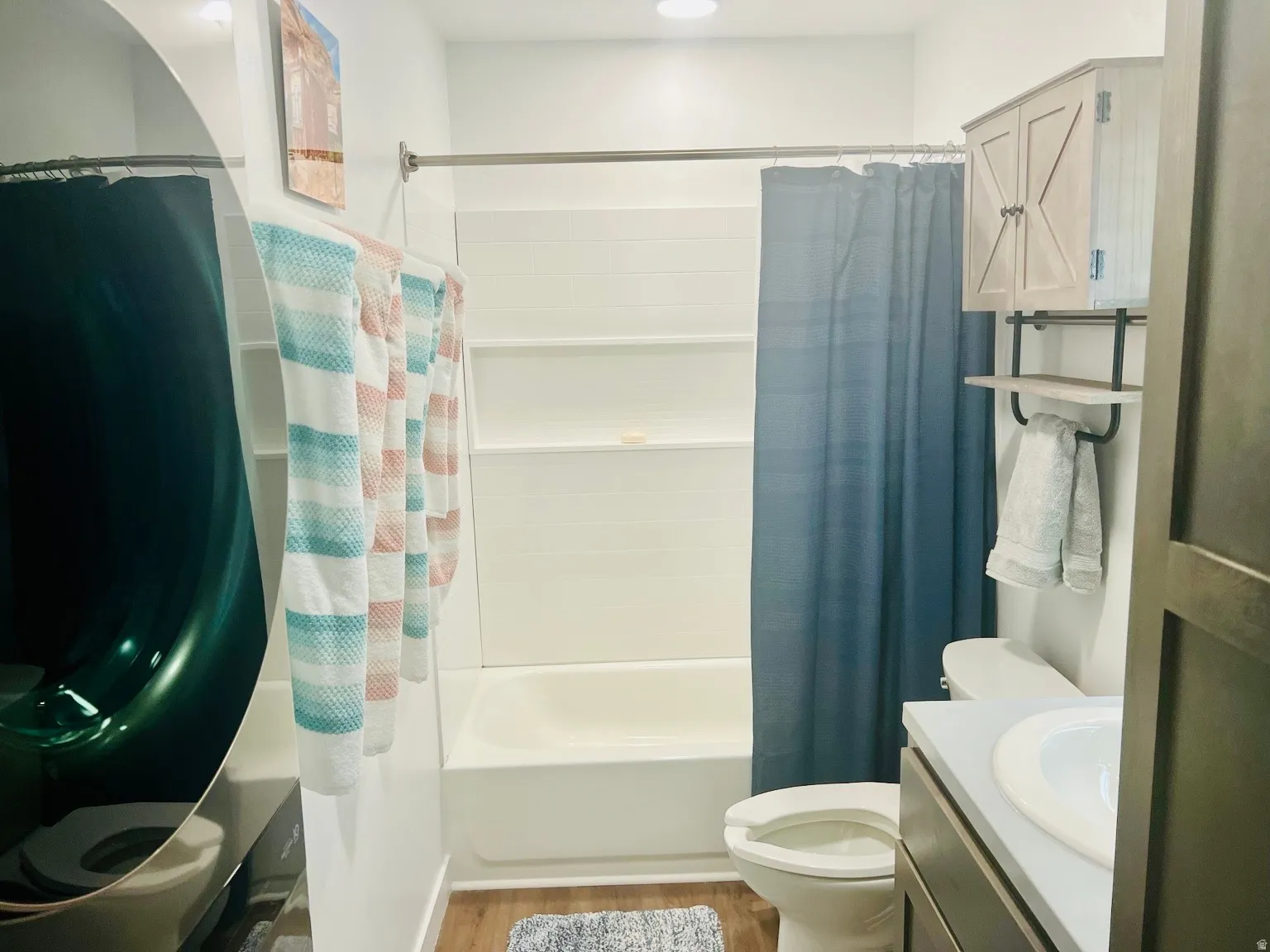 Integrated bathtub and shower with white tile surround, wood-finish vanity with a white basin sink, wall-mounted storage cabinet, and open shelving with a towel bar