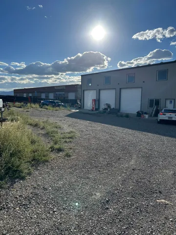 Industrial-style building with multiple roll-up garage doors, a gravel parking area, and a secondary building in the background