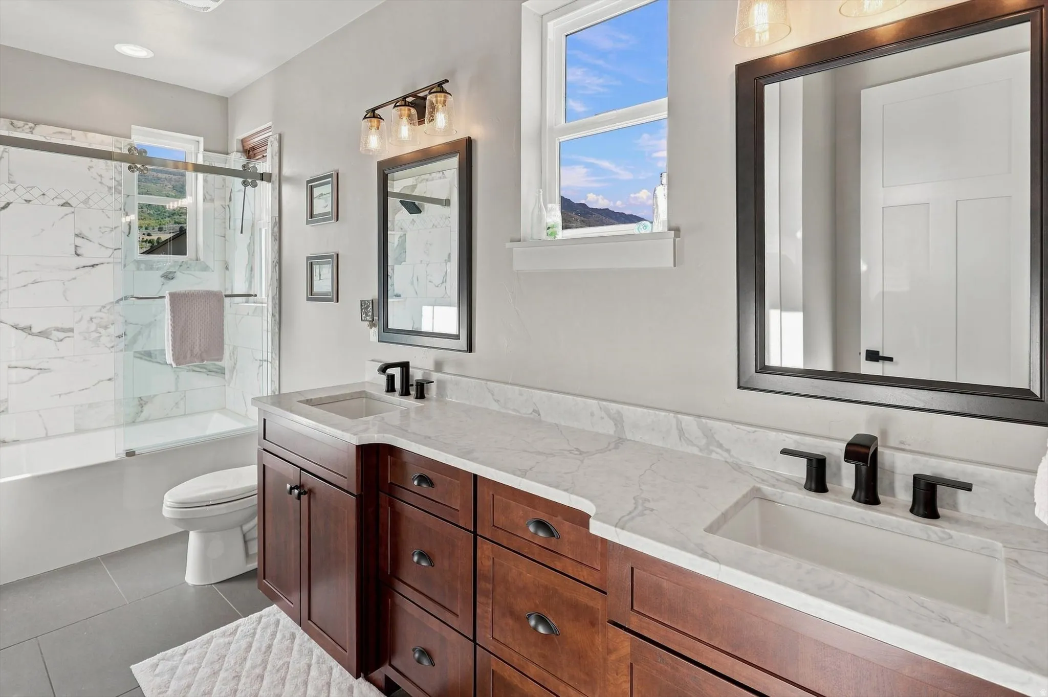 Dual sink vanity with stone-look countertop, rich wood cabinetry, matte black fixtures, and a tiled tub/shower combination with a sliding glass door