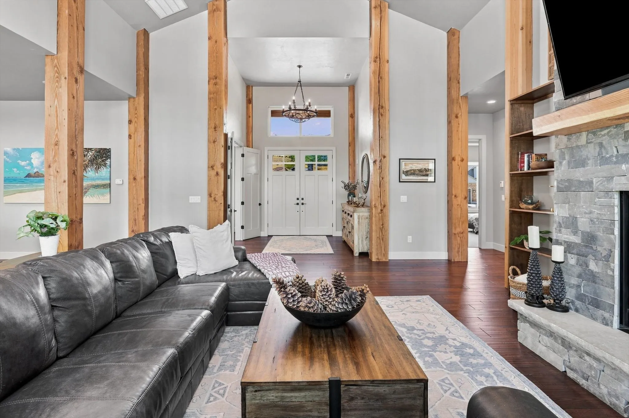Vaulted ceilings with exposed wood beams, hardwood flooring, stone fireplace with built-in shelving, white double entry doors, and a foyer chandelier